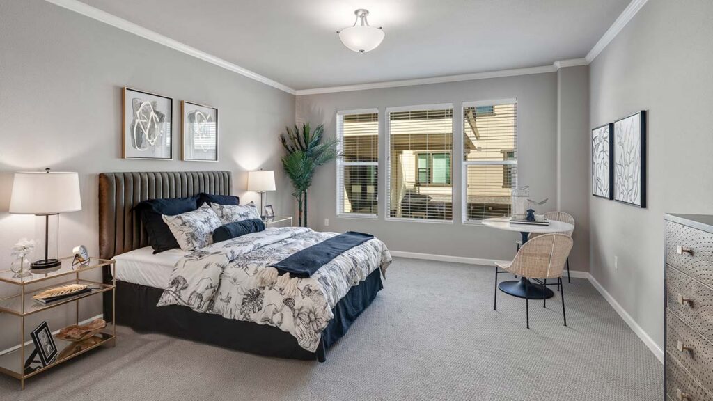 A modern bedroom with a large bed, floral bedding, two bedside tables with lamps, a small desk and chair by the window, wall art, a tall plant, and light gray walls with beige carpet. Natural light fills the room.