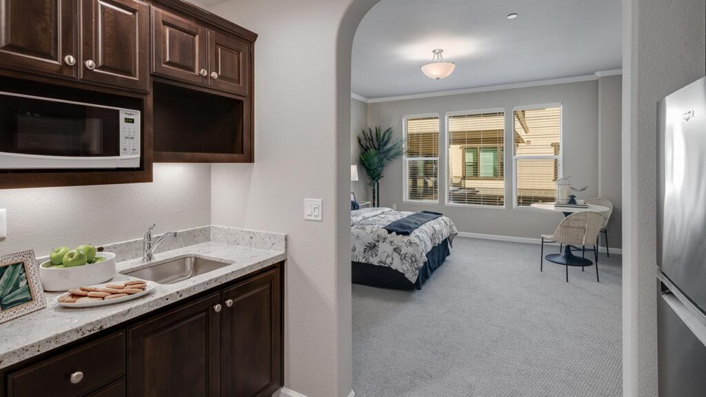 A small kitchen area with dark wood cabinets, a microwave, a sink, and a marble countertop with a plate of cookies and apples, leading into a well-lit bedroom with a bed, chair, and large windows.