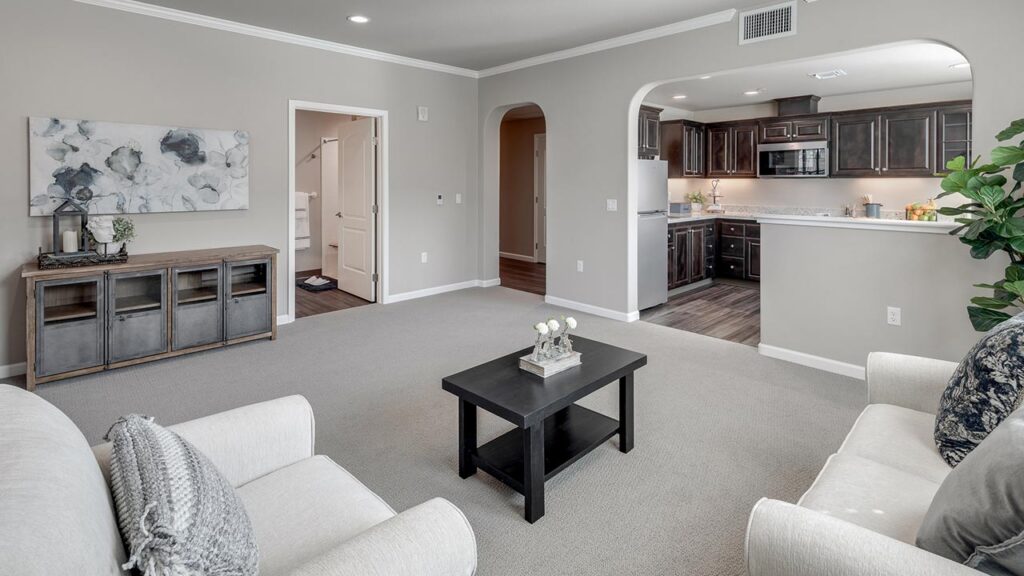 Modern living room with neutral tones, featuring two sofas, a black coffee table, and wall art. The space opens to a kitchen with dark cabinets and stainless appliances. A plant sits in the corner near a breakfast bar.