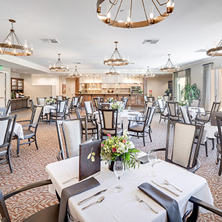 Elegant dining room with neatly set tables, floral centerpieces, upholstered chairs, and large chandeliers. The space is bright with natural light and features neutral, modern decor.
