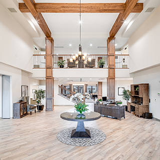 Bright, spacious lobby with wood beams, chandelier, and a round table with flowers in the center. Reception desks and office furniture are along the walls; a balcony overlooks the area above.