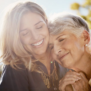A younger woman and an older woman smile warmly, embracing each other outdoors in sunlight. The older woman rests her head on the younger woman’s shoulder, and both appear happy and content.