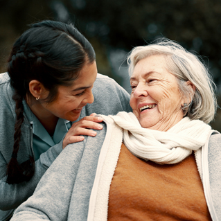 A young woman with dark hair in braids smiles warmly at an older woman with gray hair and a white scarf, who is sitting and smiling back. They appear to be enjoying a happy moment outdoors.