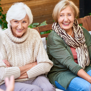 Two elderly women sit close together on a bench, smiling and laughing. One wears a beige turtleneck sweater and has arms crossed; the other wears a green cardigan with a striped scarf. They appear to be enjoying a conversation.