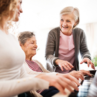 Three women of different ages are at a piano. One elderly woman is seated and playing, a younger woman sits beside her, and another older woman stands nearby, smiling and gesturing over the keys.