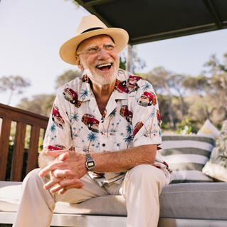 An older man wearing a straw hat, glasses, and a colorful Hawaiian shirt sits outdoors on a cushioned bench, smiling and laughing in the sunlight. Trees and a wooden railing are visible in the background.