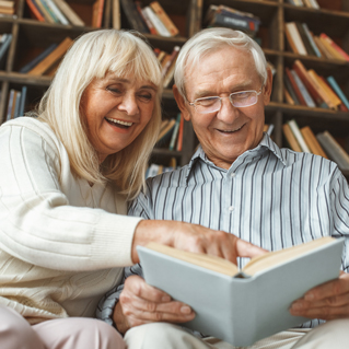 An elderly couple sits together smiling and reading a book, with bookshelves filled with books in the background. The woman points at something in the book as they enjoy their time.