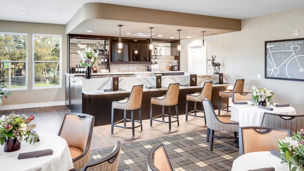 A modern dining area with round tables, floral centerpieces, and a sleek bar with leather barstools. Pendant lights hang above the bar, and large windows let in natural light. Decor includes framed art and elegant finishes.