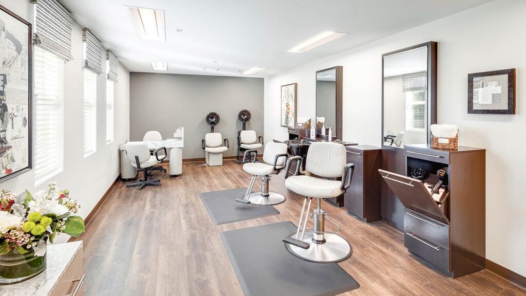 A modern, well-lit salon with wooden floors, two styling chairs at stations with mirrors, a manicure table by large windows, and two hair washing stations at the back. Fresh flowers decorate a counter on the left.