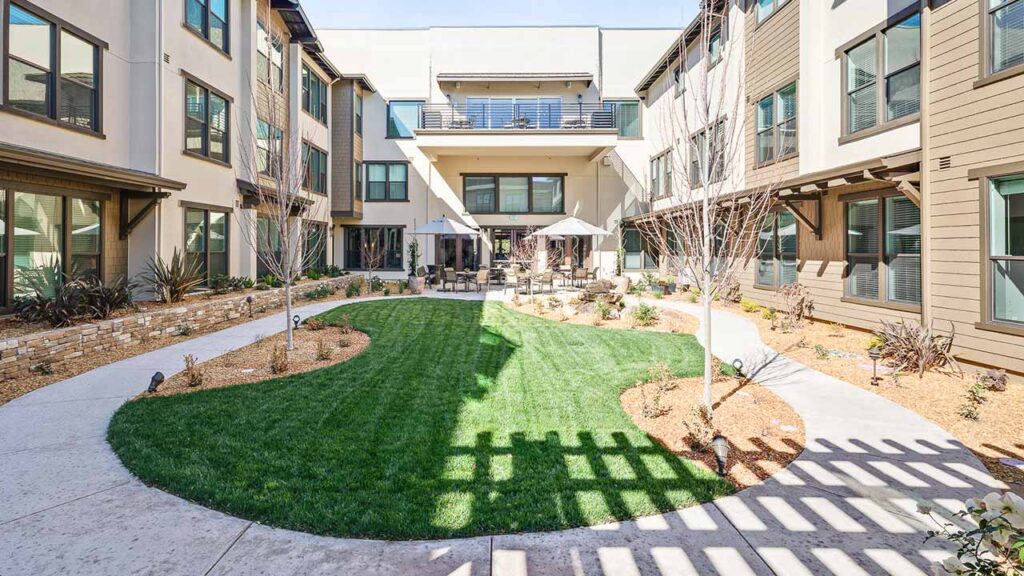 A modern apartment building with three stories surrounds a landscaped courtyard featuring green grass, small trees, outdoor seating with umbrellas, and wide walkways under a clear sky.