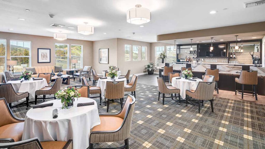 A bright, modern dining room with round tables set for meals, tan upholstered chairs, flower centerpieces, large windows, patterned carpet, and a bar area in the background.