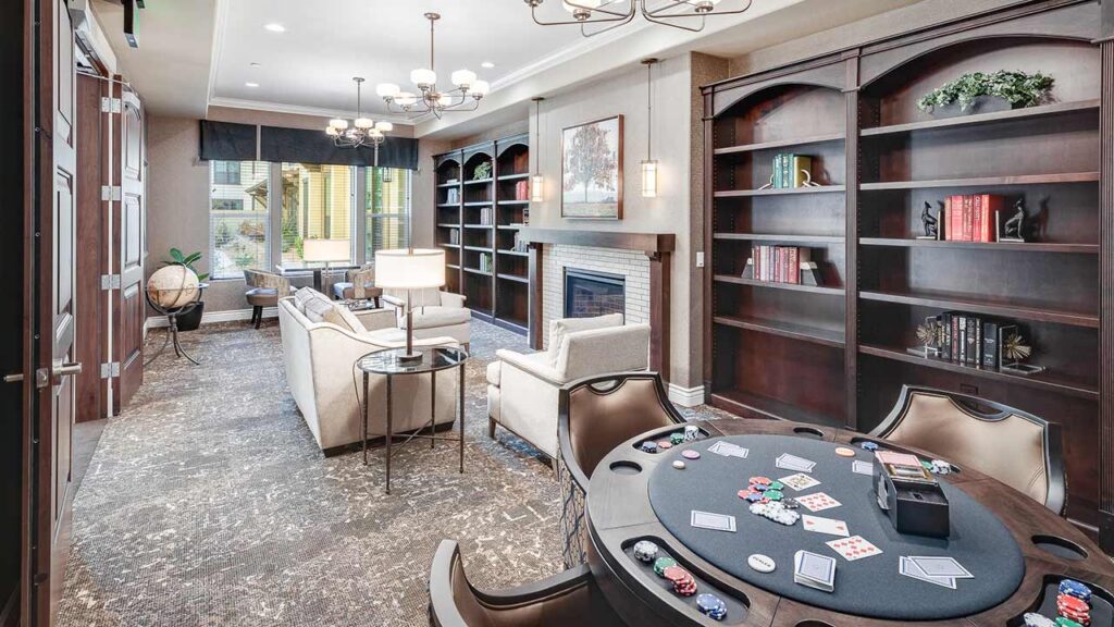 A spacious, elegant room with a poker table in the foreground, cozy sitting area with sofas near a fireplace, large bookshelves, and windows letting in natural light.