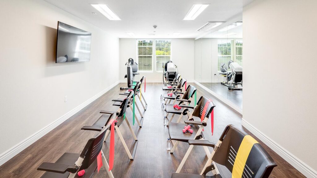 A bright exercise room with chairs in rows, each with a colored resistance band and pink dumbbells; exercise equipment is at the front, and large mirrors cover one wall.