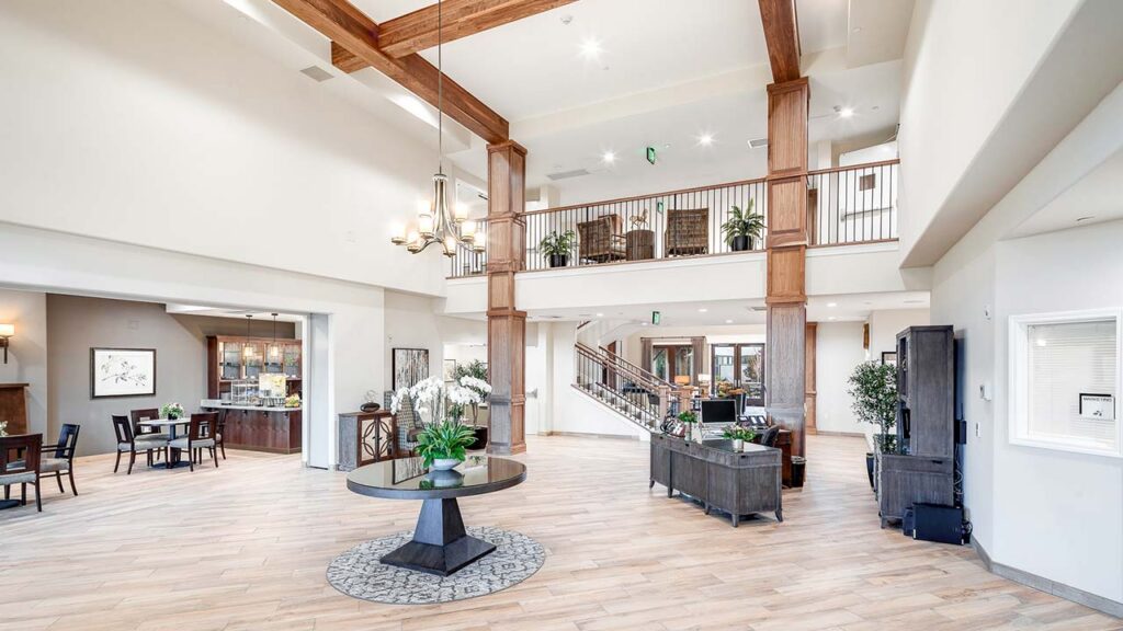 Spacious, modern lobby with high ceilings, large wooden beams, a round table with plants, and a staircase leading to an upper balcony. The area is well-lit and decorated with contemporary furniture and indoor plants.
