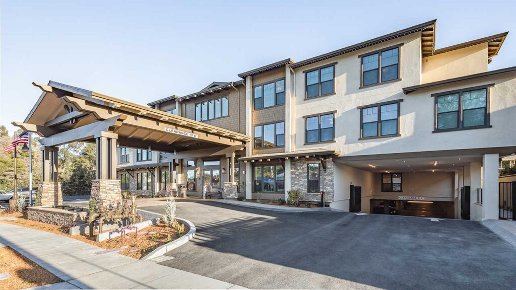 Modern three-story building with large covered entrance, stone pillars, and American flag. Driveway leads to main entry and an underground parking garage. Landscaping with rocks and mulch borders the building.