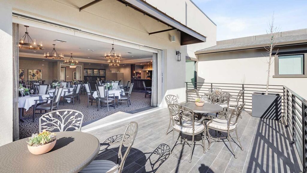 A modern dining area with large glass doors opens onto an outdoor patio with metal tables and chairs, potted succulents, and a railing, allowing natural light to fill both spaces.