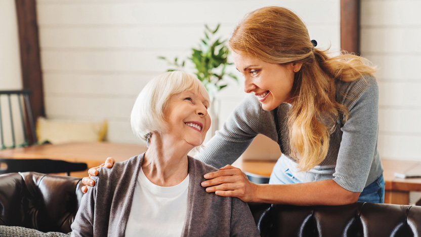A younger woman smiles and leans over to gently touch the shoulders of an older woman, who is seated on a couch and smiling back—both enjoying a warm moment together during memory care activities indoors.