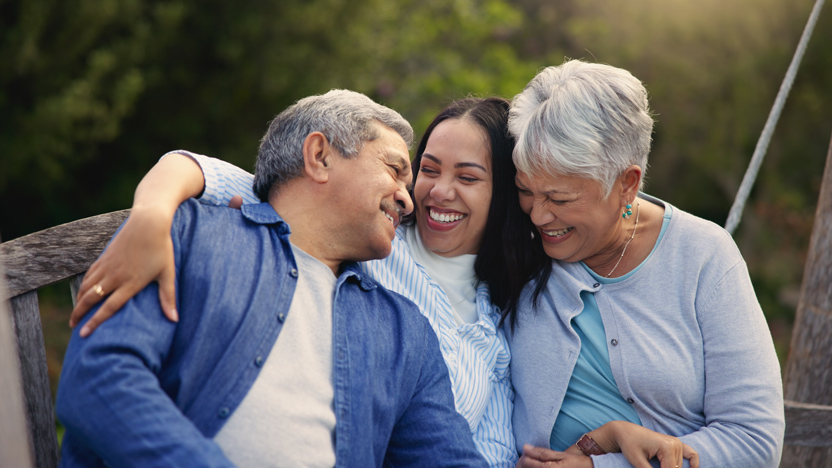 Three people, two older adults and a younger woman, sit closely together on a bench outdoors, smiling and laughing, with greenery in the background. The mood is warm and joyful.